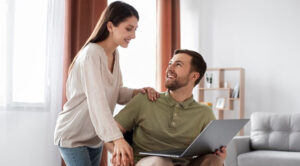 A woman rests her hand on the shoulder of a smiling man holding a laptop at home