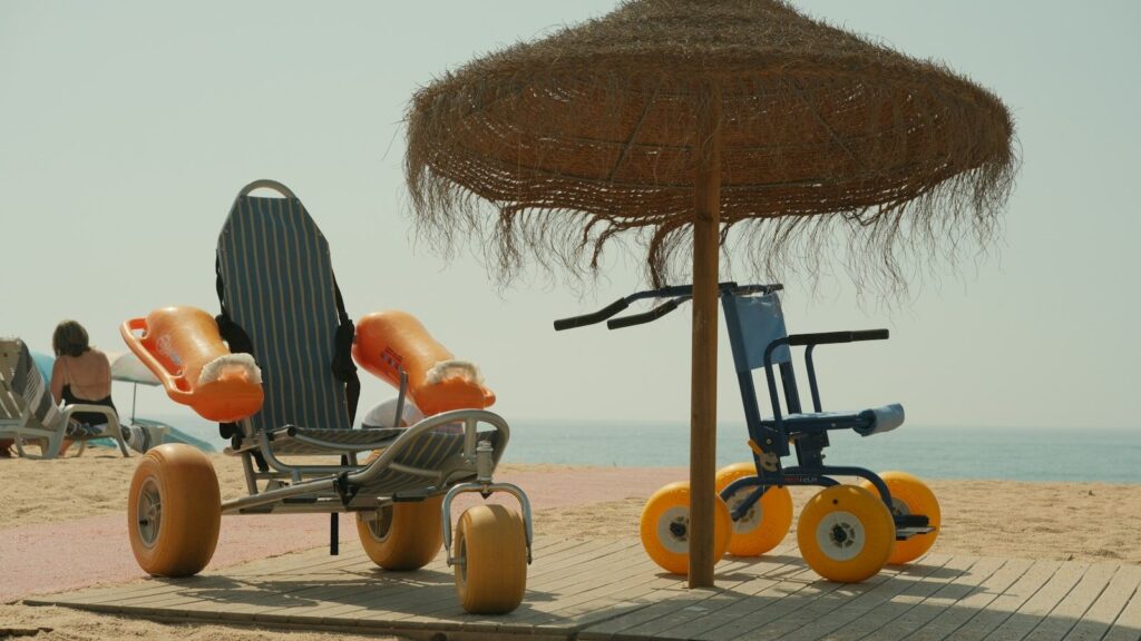 Two beach wheelchairs with large sand wheels under a straw umbrella on the sand